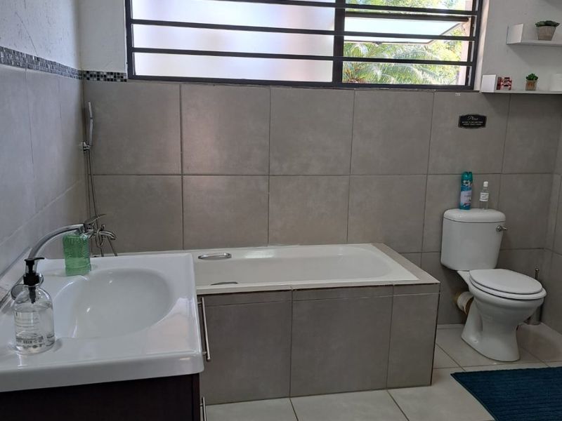A functional bathroom featuring a built-in bathtub, vanity basin, and toilet, finished with neutral wall and floor tiling and a horizontal window.