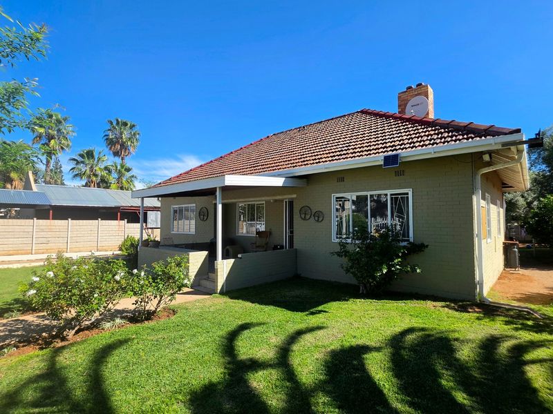Rear view of a single-story brick house featuring a tiled roof, chimney, and a covered patio area overlooking a well-maintained garden.