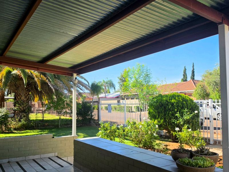 A covered outdoor patio area featuring a corrugated metal roof, steel support beams, and a low brick wall with a tiled ledge overlooking the garden.