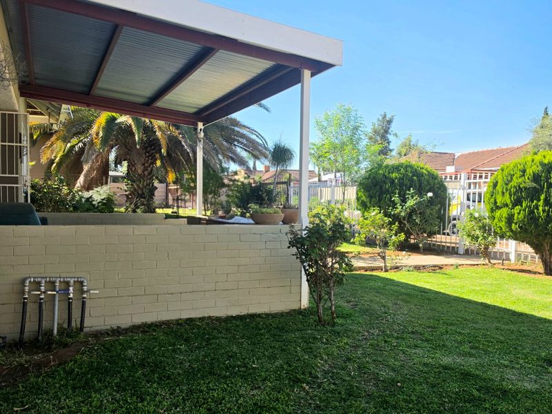 A covered outdoor patio area overlooking a well-maintained garden, featuring a brick wall and exposed exterior plumbing fixtures.