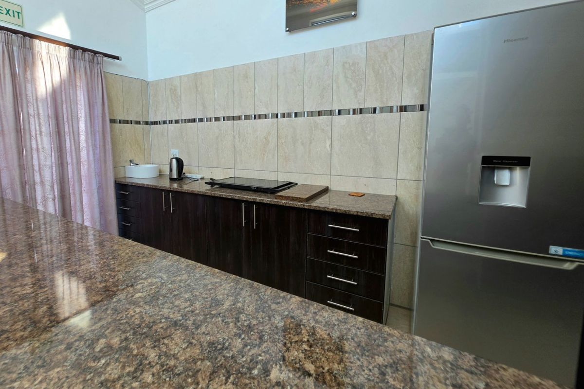 A functional kitchen featuring dark wood cabinetry, granite countertops, a tiled backsplash, and a built-in hob with an integrated stainless steel refrigerator.