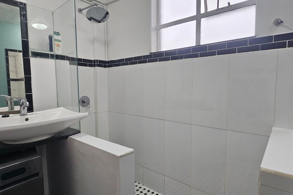 A functional bathroom featuring a walk-in shower with glass screen, basin, and toilet, finished with white wall tiles and a dark accent border.