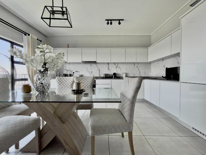 Modern open-plan kitchen and dining area featuring sleek white handleless cabinetry, marble-look splashback, tiled floors, and contemporary lighting fixtures.