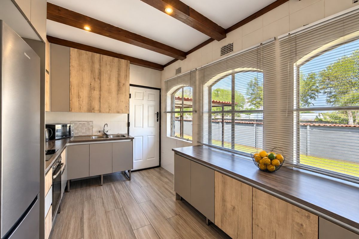 Modern kitchen featuring wood-look cabinetry, exposed timber ceiling beams, tiled flooring, and large arched windows providing ample natural light.