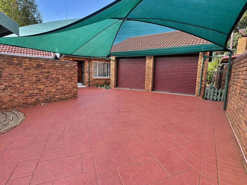 A well-maintained front exterior featuring a paved driveway, double garage with roll-up doors, face brick construction, and a large protective shade sail.