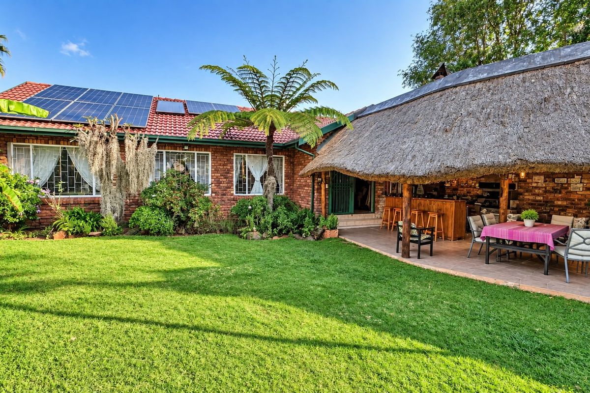 Spacious rear garden featuring a well-maintained lawn, a covered thatched patio with a built-in bar, and a brick house equipped with solar panels.