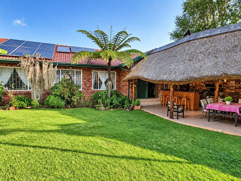 Spacious rear garden featuring a well-maintained lawn, a covered thatched patio with a built-in bar, and a brick house equipped with solar panels.
