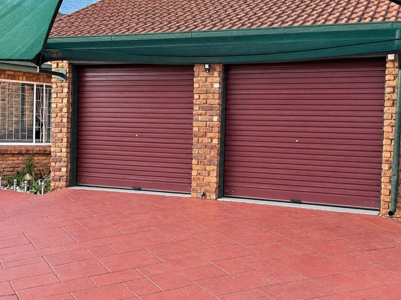 A well-maintained double garage featuring red roller doors, face brick exterior, and a paved driveway area covered by a shade cloth canopy.
