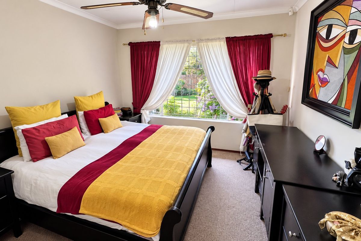 A well-maintained bedroom featuring a ceiling fan, carpeted flooring, and a window with security bars overlooking the garden.