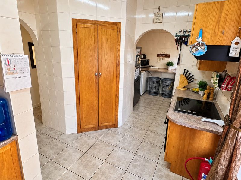 A functional kitchen featuring tiled walls and flooring, wooden cabinetry, and an electric cooktop, with an adjacent scullery area accessible through an archway.