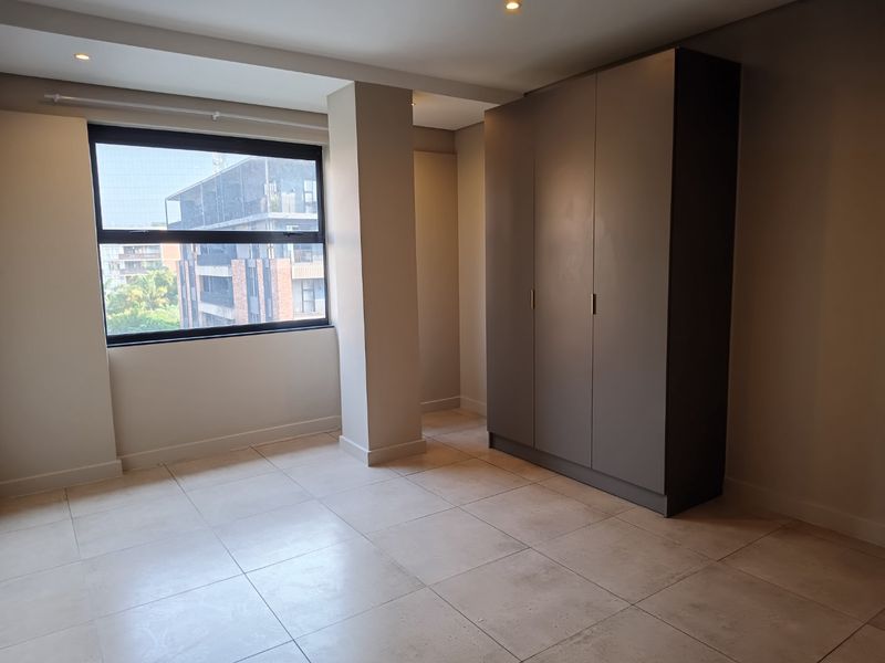 A bright, empty bedroom featuring large format floor tiles, recessed lighting, and a modern built-in wardrobe with a neutral finish.
