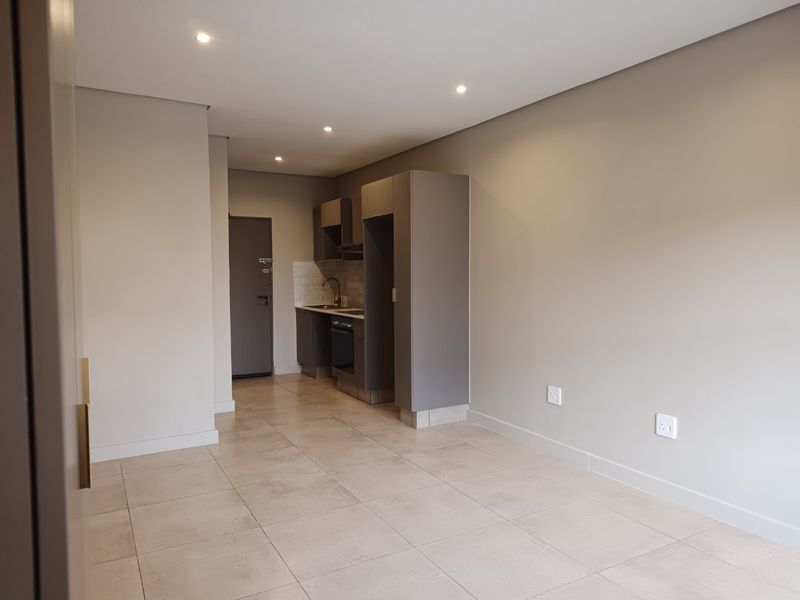 Modern open-plan living area featuring a compact kitchen with built-in cabinetry, oven, and hob, finished with neutral tiled flooring and recessed lighting.