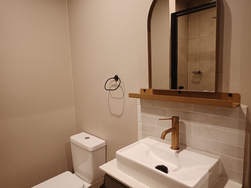 Modern bathroom featuring a vanity with a brass faucet, tiled backsplash, arched mirror with integrated shelf, and a standard toilet.