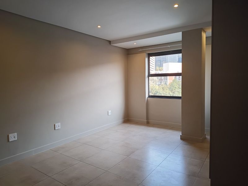 A bright, empty bedroom featuring neutral tiled flooring, recessed ceiling lighting, and a window with an urban view.
