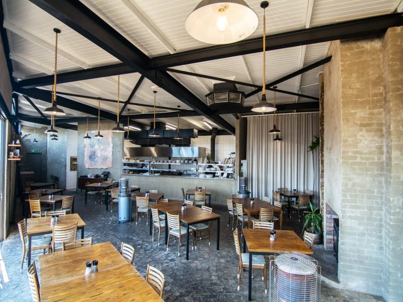 Spacious open-plan dining area featuring exposed steel ceiling trusses, industrial pendant lighting, a brick fireplace, and a functional commercial kitchen pass.