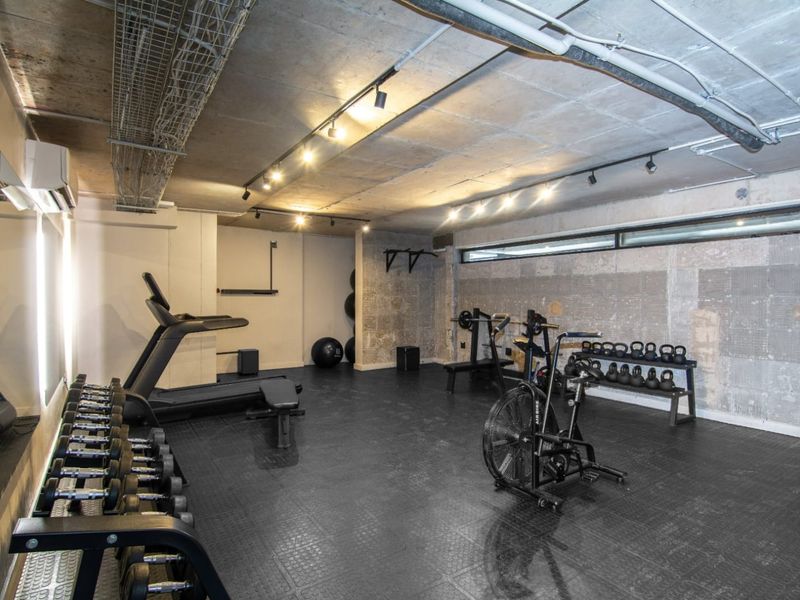 A modern industrial-style gym featuring exposed concrete ceilings, track lighting, durable rubber flooring, and a large wall-mounted mirror.