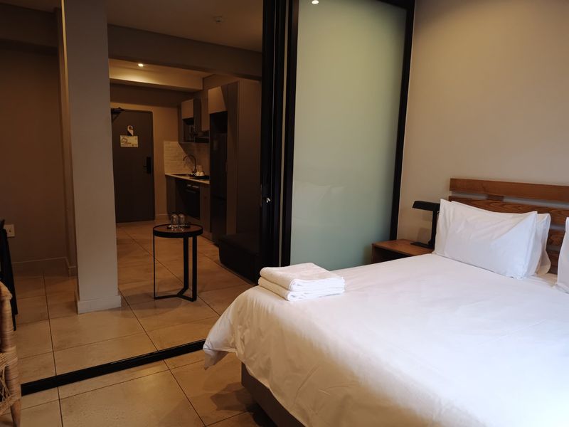 An open-plan apartment interior featuring a bedroom area separated by a frosted glass partition from a compact, modern kitchen with built-in cabinetry.