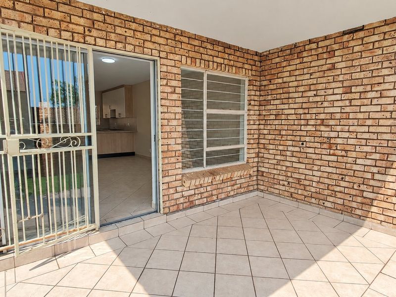 A covered patio area featuring exposed brick walls, tiled flooring, and a secure sliding door entrance leading into the home.