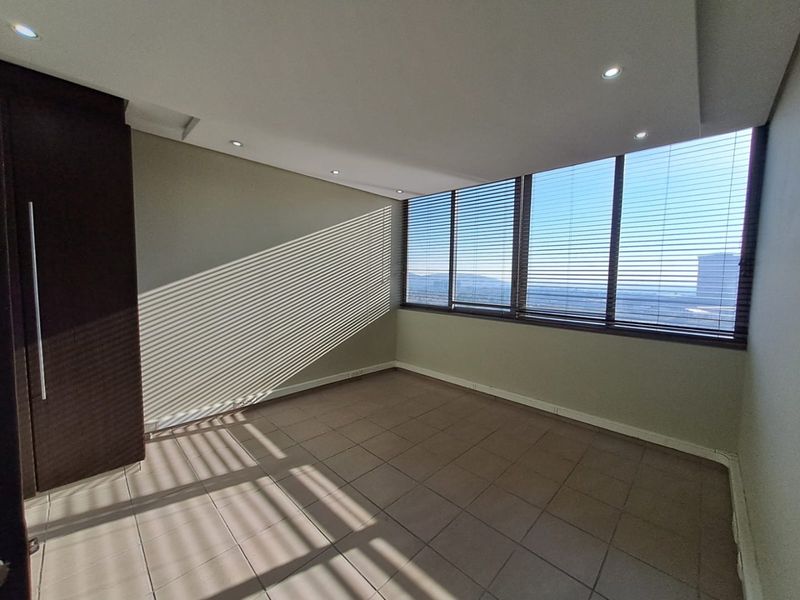 A bright, empty bedroom featuring tiled flooring, built-in cupboards, recessed downlighting, and large windows with venetian blinds offering expansive views.