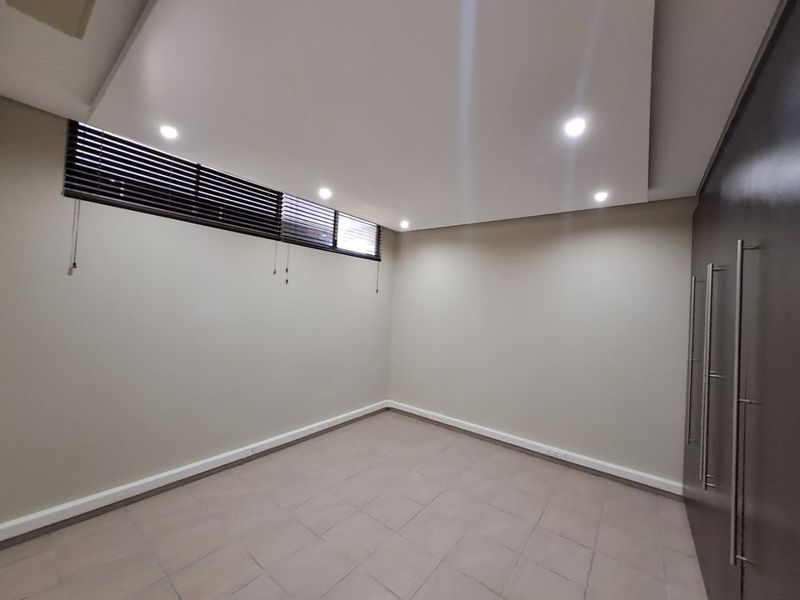 A clean, empty bedroom featuring tiled flooring, recessed ceiling lighting, built-in cupboards, and horizontal venetian blinds.