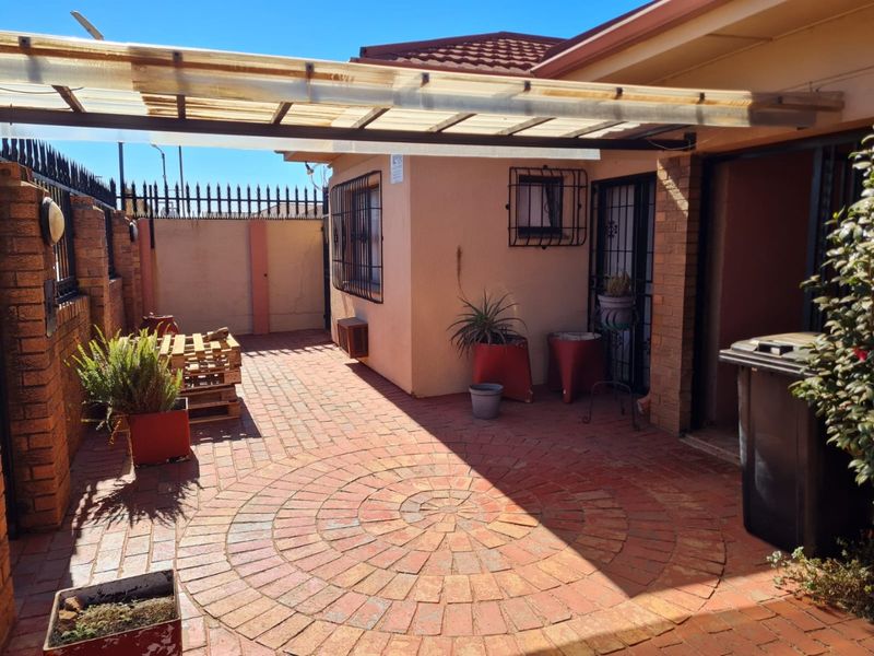 A paved outdoor patio area featuring a covered roof structure, brick boundary walls, and security fixtures, providing a functional space for outdoor living.