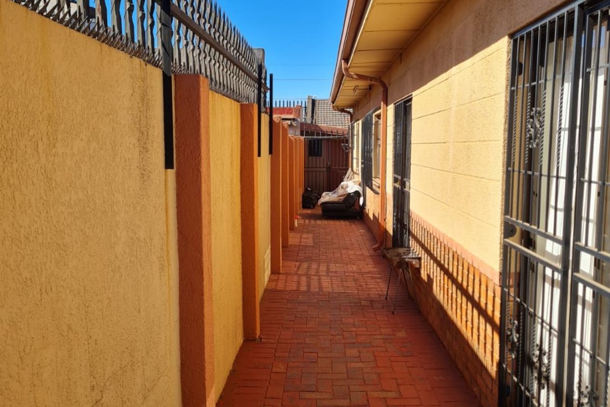 A narrow side passage featuring a paved walkway, high boundary walls with security spikes, and secure metal gates and window bars.
