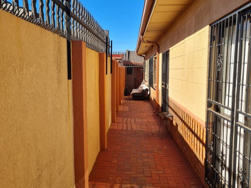 A narrow side passage featuring a paved walkway, high boundary walls with security spikes, and secure metal gates and window bars.