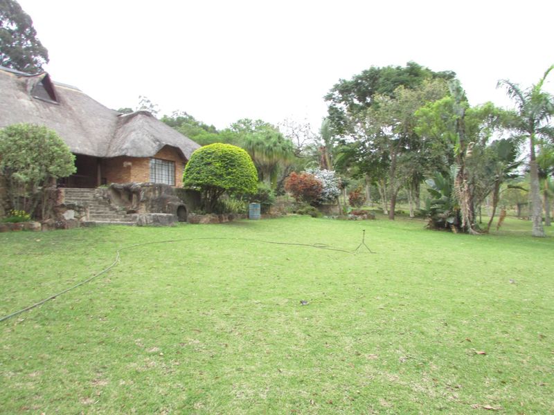 A spacious rear garden featuring a well-maintained lawn, mature trees, and a charming farmhouse with a traditional thatch roof and stone entrance steps.