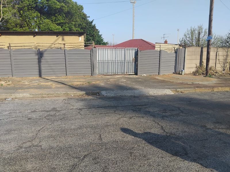 Front exterior view showing a gated entrance with a precast concrete boundary wall and paved driveway access from the street.