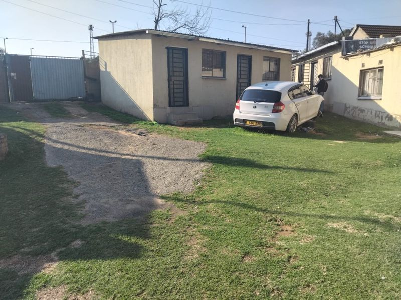 Exterior view of a residential property featuring a gravel driveway, security gates, and basic building structure with visible wear to the exterior walls.
