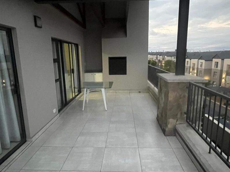 A spacious covered balcony featuring tiled flooring, a built-in braai, and metal railings, offering an ideal outdoor entertainment space.