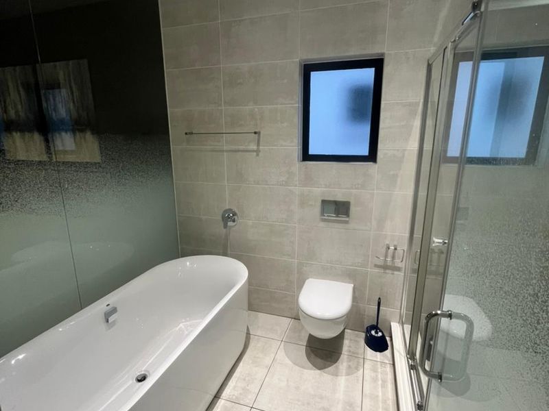 Modern bathroom featuring a sleek freestanding bathtub, wall-mounted toilet, glass-enclosed shower, and neutral floor-to-ceiling tiling with recessed lighting.