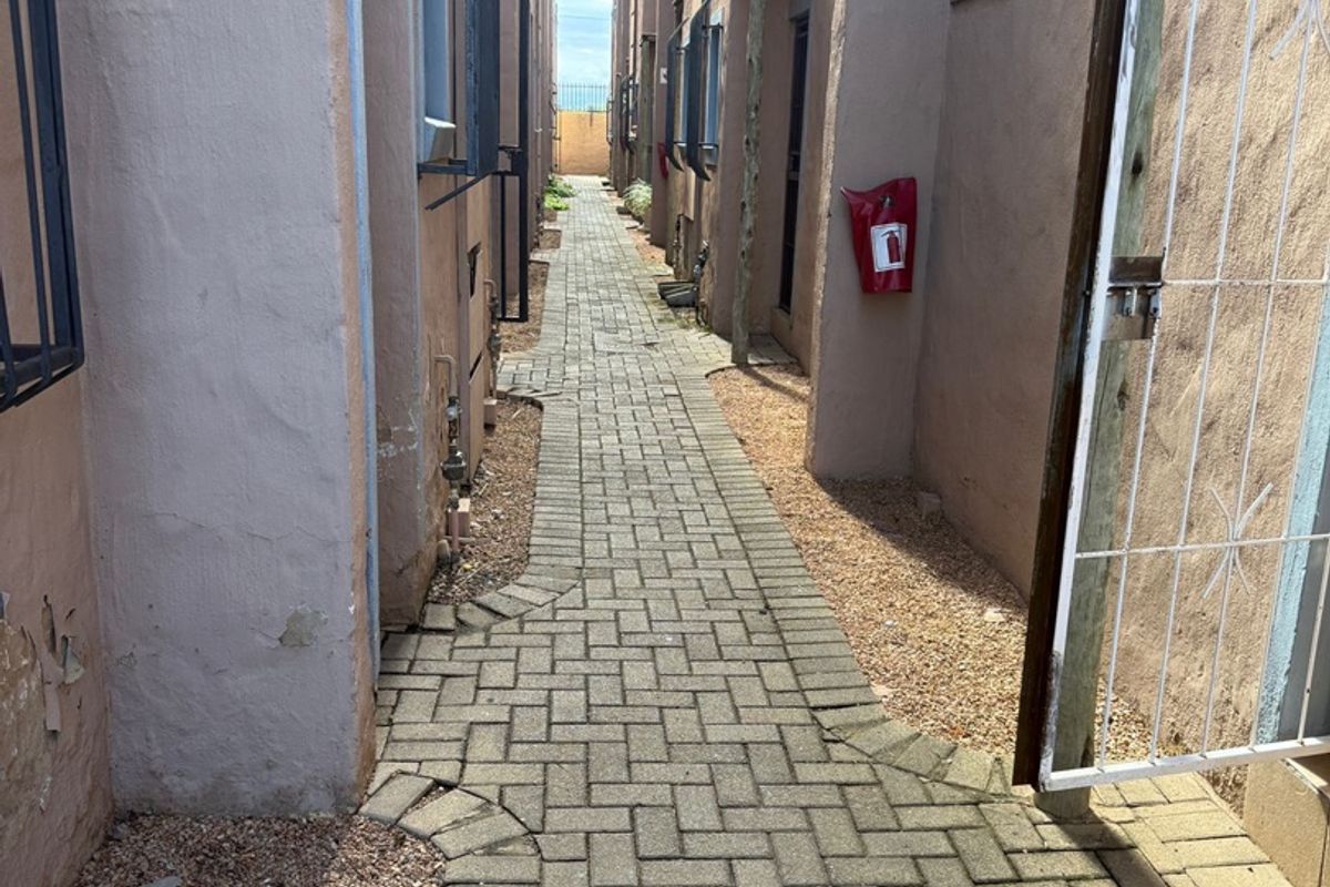 A narrow paved walkway between apartment building blocks featuring security gates, fire safety equipment, and exterior wall access.