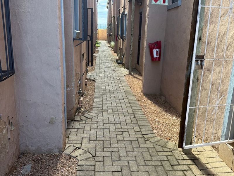 A narrow paved walkway between apartment building blocks featuring security gates, fire safety equipment, and exterior wall access.