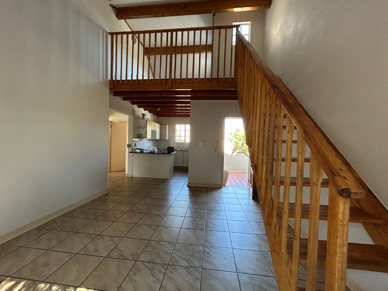 Open-plan living area featuring tiled floors, a wooden staircase leading to a mezzanine level, exposed ceiling beams, and an adjoining kitchen space.