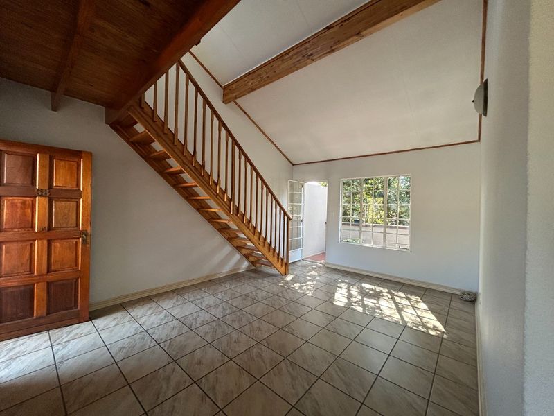 Bright, open-plan living area featuring a wooden staircase, exposed ceiling beams, and tiled flooring with natural light from a grid window.