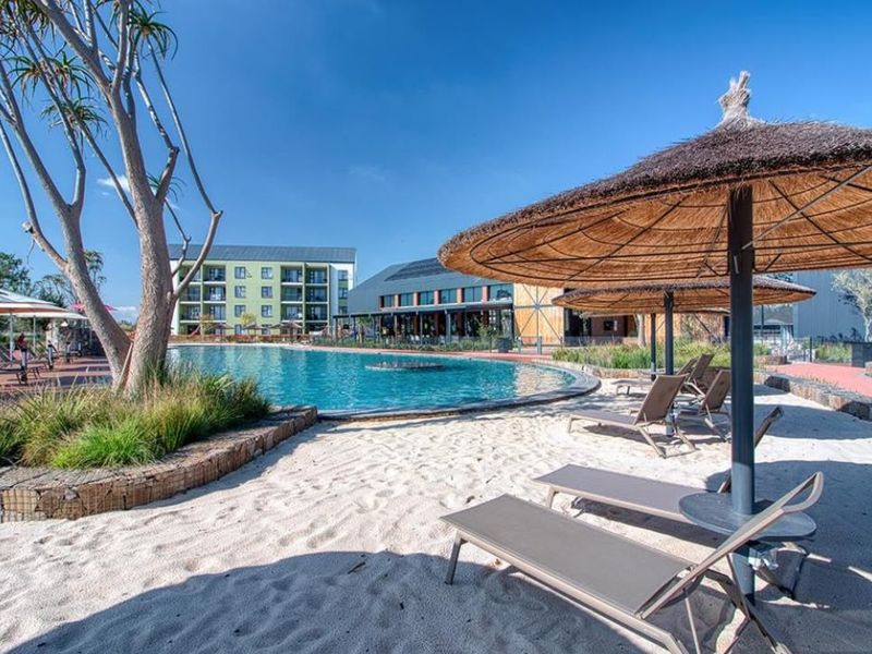 A well-maintained outdoor swimming pool area featuring a sandy beach section, landscaped surroundings, and paved walkways within a modern apartment complex.