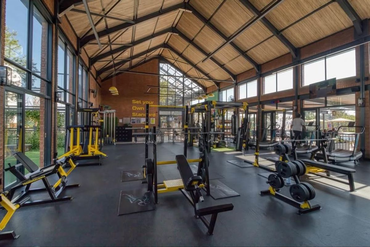 A spacious, modern gym facility featuring high vaulted ceilings with exposed steel trusses, large glass windows, and durable rubber flooring.