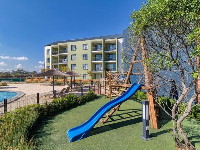 Communal outdoor area featuring a swimming pool, sandy beach area, and a children's playground set against the apartment building facade.