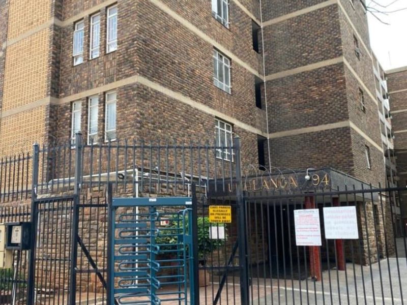 Front exterior view of a multi-story brick apartment building featuring secure gated access, a turnstile, and perimeter security fencing.