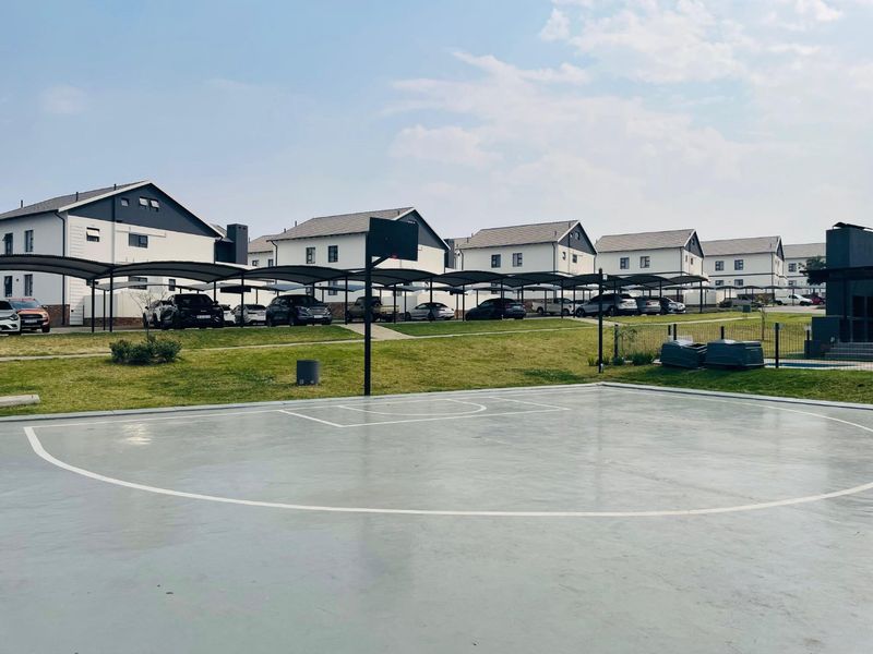 A well-maintained outdoor basketball court situated within a residential townhouse complex, featuring paved surfaces and adjacent covered parking bays.