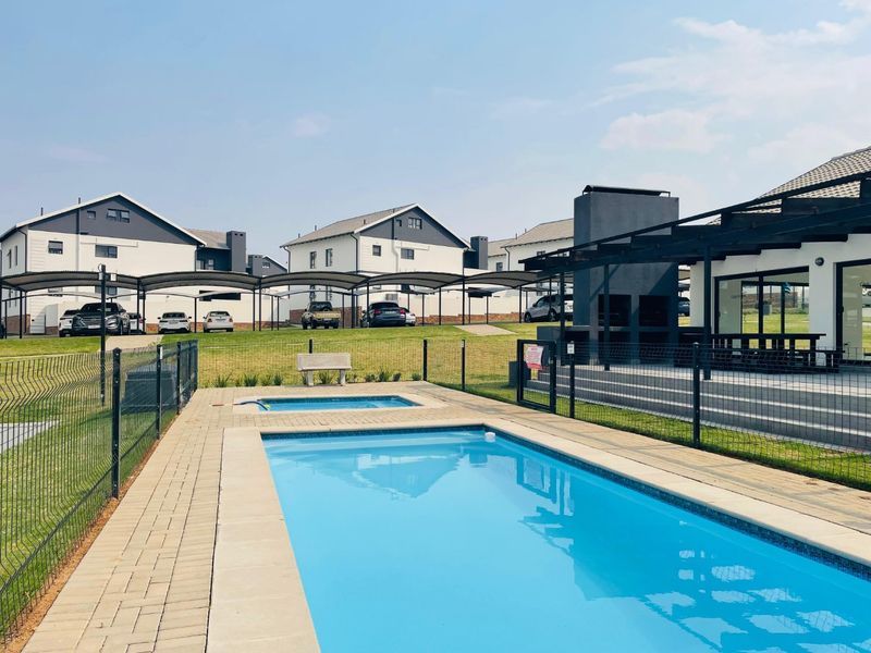 Communal pool area featuring a swimming pool, paved surround, and a covered patio with a built-in braai, set within a modern townhouse complex.