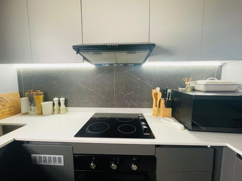 Modern kitchen featuring sleek grey cabinetry, a dark stone-look backsplash, integrated electric hob, oven, and extractor fan with contemporary under-cabinet lighting.