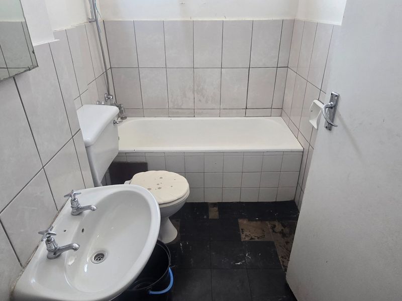 A dated bathroom featuring a standard bathtub, pedestal basin, and toilet with worn floor and wall tiling, requiring renovation to modernize the space.
