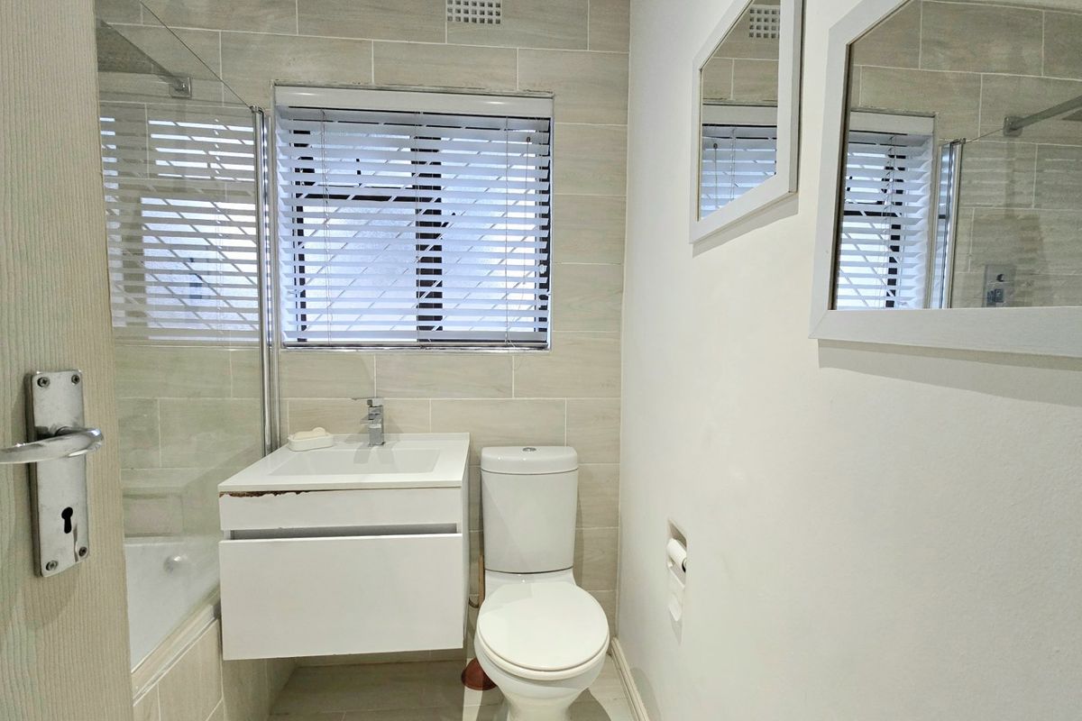 A compact bathroom featuring a wall-mounted vanity, toilet, and a bathtub with a glass shower screen, finished with neutral wall and floor tiling.