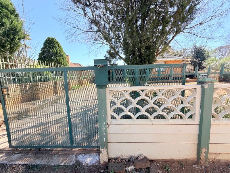 Front entrance view featuring a metal security gate, decorative concrete screen wall, and established garden trees.