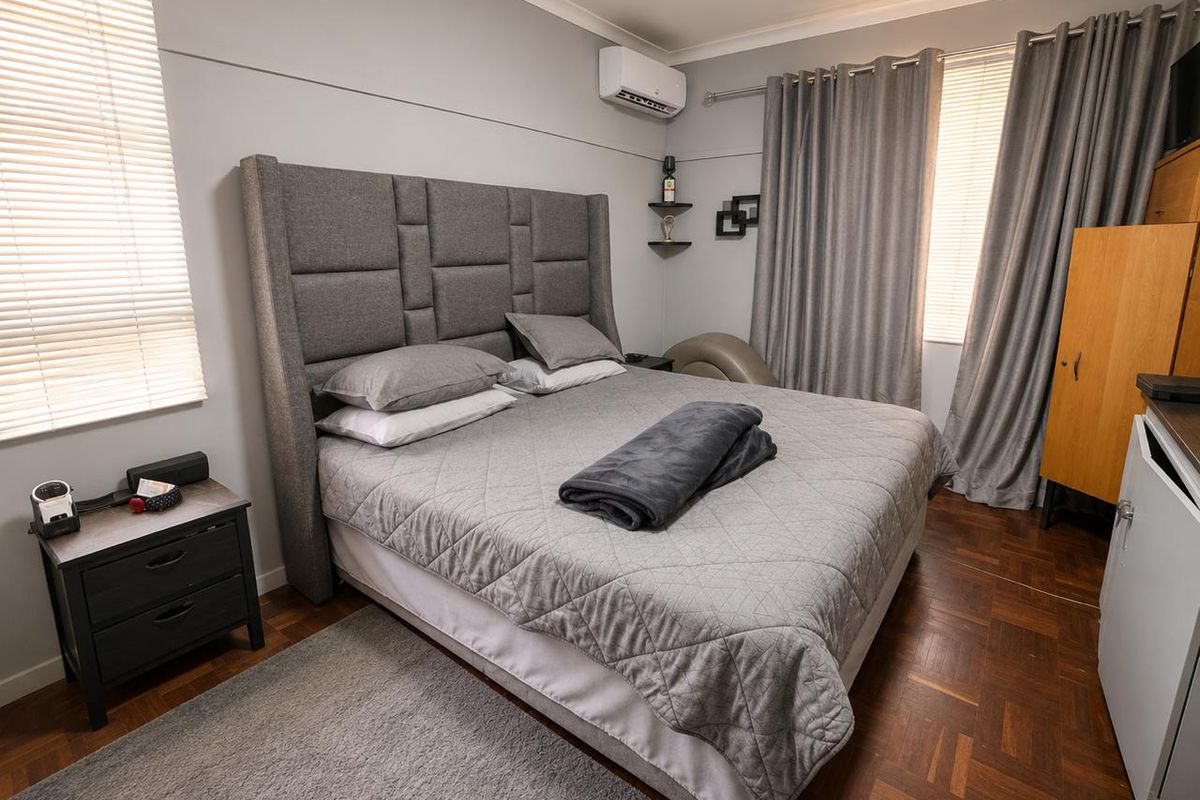 A well-maintained bedroom featuring classic parquet flooring, a wall-mounted air conditioning unit, and neutral wall finishes with dual window treatments.