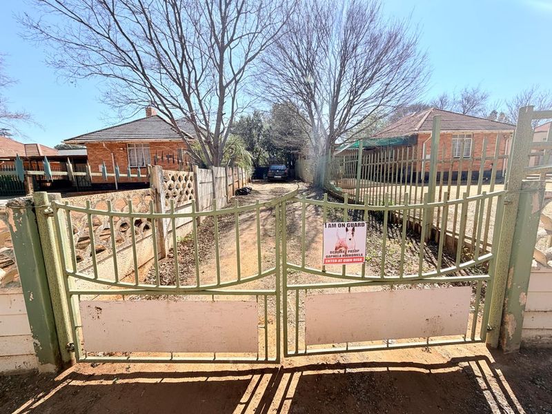 Front view of a residential property featuring a metal driveway gate, brick facade, and tiled roof, with a gravel driveway leading to the house.