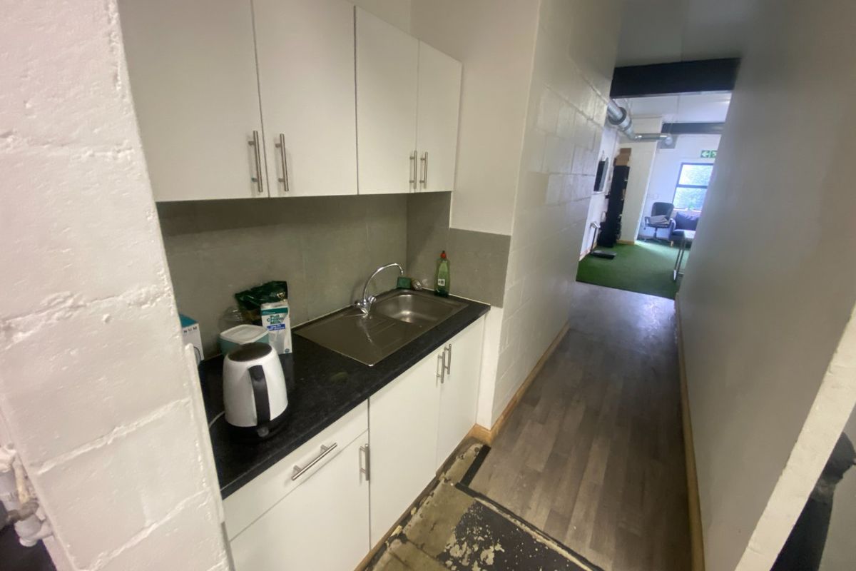 Compact kitchenette area featuring white cabinetry, stainless steel sink, and laminate countertops, leading into an open-plan commercial space with exposed industrial ducting.