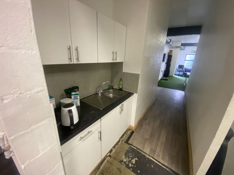 Compact kitchenette area featuring white cabinetry, stainless steel sink, and laminate countertops, leading into an open-plan commercial space with exposed industrial ducting.